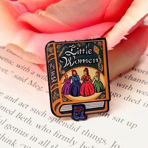 May include: A rectangular pin shaped like a book with the title "Little Women" on the cover. The pin features illustrations of women in colorful dresses. A rose bookmark is attached. The pin is set against a backdrop of a pink rose and book pages.
