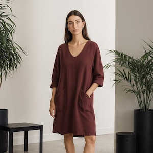 Può includere: Abito bordeaux con scollo a V, maniche a tre quarti e due ampie tasche frontali. L'abito è lungo fino al ginocchio e indossato con zoccoli di cuoio neri. L'ambientazione è una stanza minimalista con piante in vaso e mobili neri.