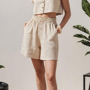 May include: Beige linen two-piece set, featuring a short-sleeved, button-down shirt and elastic-waist shorts. The outfit is paired with orange sandals. The ensemble is suitable for warm weather and casual occasions.