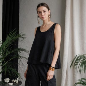 Può includere: Un top nero senza maniche e pantaloncini cargo abbinati. L'outfit è realizzato in un tessuto leggero. La modella indossa orecchini d'oro e un braccialetto di legno. Lo sfondo comprende piante e uno sfondo di colore neutro.