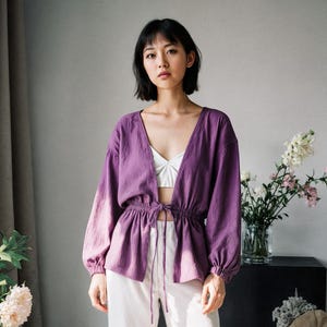 Puede incluir: Una persona con una blusa morada de manga larga con cuello en V y cintura con cordón, combinada con pantalones blancos y sandalias negras. La blusa tiene un efecto degradado, que pasa del morado a un tono más claro. Flores y una mesa negra en el fondo.