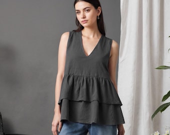 May include: A dark charcoal gray sleeveless top featuring a V-neckline and two tiered ruffled layers at the hem, paired with light blue denim jeans and brown leather sandals. The top has a relaxed fit and a solid, matte fabric texture.