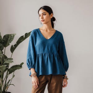 May include: A woman in a teal blue V-neck top with long sleeves and brown trousers. The top has a gathered waist. The trousers are wide-legged. The woman is wearing sandals. A large potted plant is to the left.