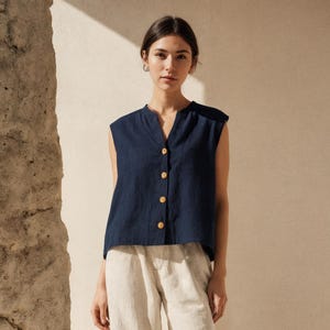 May include: A sleeveless navy blue button-down top with a V-neckline and light-coloured buttons, paired with loose-fitting, ankle-length beige trousers and dark brown leather slip-on shoes.