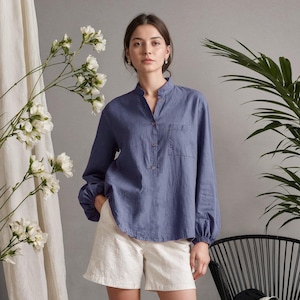 Puede incluir: Una mujer con una camisa de manga larga azul bígaro con un bolsillo y pantalones cortos blancos. La camisa tiene cuello mandarín y cierre de botones. Lleva zapatos blancos. El fondo incluye plantas y una silla negra.