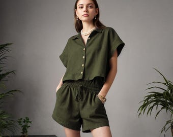 May include: An olive green two-piece set consisting of a short-sleeved button-down collared shirt and matching high-waisted shorts with an elastic drawstring waistband and side pockets, paired with bright orange slide sandals.