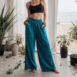 Puede incluir: Una mujer con un top corto negro y pantalones anchos color teal. Los pantalones tienen bolsillos y un corte holgado. Lleva pendientes y pulseras de madera. El fondo incluye plantas, una ventana y una vista del océano.