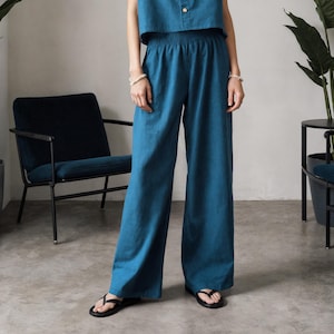 May include: Teal-colored two-piece outfit. The top is a short-sleeved button-down shirt with a collar. The high-waisted pants are wide-legged. The outfit is made of a soft fabric. The model is wearing black sandals and a pearl necklace.