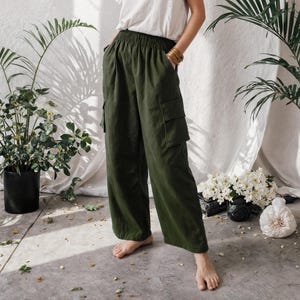 May include: A person wearing a white t-shirt and olive green cargo trousers. The trousers feature large side pockets and a relaxed fit. The outfit is accessorised with gold hoop earrings and bracelets. The background includes plants and a white curtain.