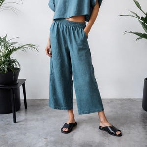 May include: A teal two-piece linen outfit. The set includes a short-sleeved crop top and wide-leg pants with an elastic waistband. The model is wearing black sandals. The outfit is displayed in a bright, minimalist setting with potted plants.