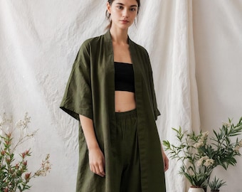 May include: An olive green linen outfit consisting of a long, open-front kimono-style jacket, matching cropped pants, and a black tube top. The model is wearing black sandals. The outfit is displayed in a studio setting with plants.