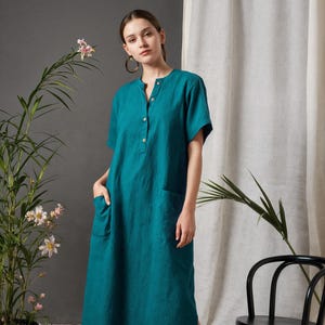 Può includere: Abito in lino color teal con maniche corte, colletto alla coreana e dettagli con bottoni. L'abito ha due tasche frontali e uno spacco laterale. La modella indossa ballerine nere. Una sedia nera e piante in vaso sono sullo sfondo.