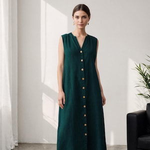 May include: A sleeveless, dark green dress with a V-neck and a button-down front. The dress falls to the mid-calf and is worn with black loafers. The dress is made of a soft, flowing fabric.