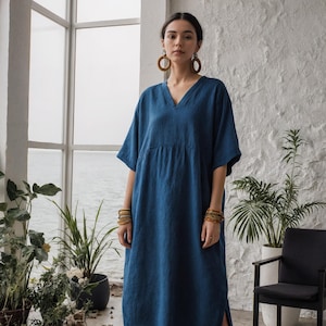 May include: A long, flowing, indigo blue dress with a V-neck and short sleeves. The dress has a relaxed fit and a side slit. The model is wearing wooden hoop earrings and multiple gold bracelets. The background includes plants and a window.