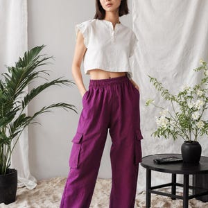 May include: A woman in a white crop top with frilled sleeves and purple cargo trousers. The trousers have large side pockets. She wears black sandals. A black side table and a potted plant are in the background.