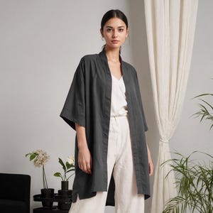Puede incluir: Una chaqueta estilo kimono gris carbón con mangas anchas, usada sobre una camiseta blanca y pantalones de pierna ancha color crema. El atuendo es parte de una estética minimalista y moderna. El fondo incluye una cortina blanca y plantas en macetas.