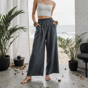 May include: A woman wearing a white crop top and wide-leg, dark gray pants. The pants have an elastic waistband and pockets. The background includes plants, a beach scene, and a white wall.