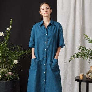 May include: A teal-colored linen shirt dress with short sleeves, a button-down front, and two front pockets. The dress falls to the mid-calf and is paired with black leather loafers. The background includes plants and a black table.