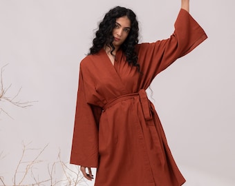 May include: A rust-colored wrap robe featuring wide, long sleeves and a matching fabric tie belt at the waist. The garment has a V-neckline and falls to a mid-thigh length, paired with brown strappy sandals.