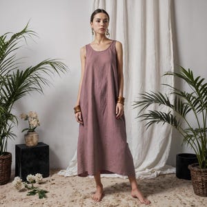 Op de afbeelding: Een mouwloze, stoffig roze jurk met een ronde hals. De jurk is enkellang en gemaakt van een zachte, vloeiende stof. Het model draagt houten armbanden en oorbellen. De achtergrond omvat potplanten en een wit gordijn.