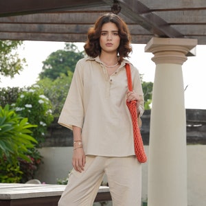 May include: A woman wearing a beige linen shirt and trousers with a coral crochet bag. She is standing in front of a wooden pergola and a white pillar.