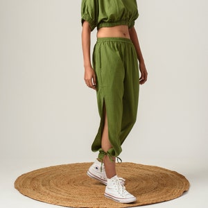 Puede incluir: Top corto verde oliva con mangas cortas abullonadas y pantalones a juego. Los pantalones tienen una abertura lateral y están atados en los tobillos. El conjunto se combina con zapatillas altas blancas. La modelo está de pie sobre una alfombra redonda tejida.