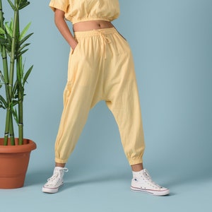 May include: A pale yellow two-piece set consisting of a short-sleeved cropped top with a round neckline and matching high-waisted harem-style trousers featuring an elasticated drawstring waistband and cuffed ankles, paired with white high-top trainers.