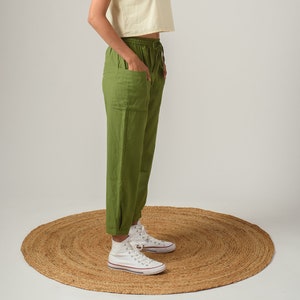 May include: A woman wearing a light yellow cropped t-shirt and olive green pants with a drawstring waist. She is wearing white high-top sneakers and standing on a woven rug.