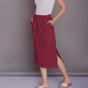 Könnte beinhalten: Eine Frau trägt ein weißes Tanktop und einen maroonfarbenen Midirock mit einem seitlichen Schlitz. Der Rock hat einen elastischen Bund und Taschen. Sie trägt weiße Sandalen.
