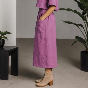 May include: A woman in a lilac linen co-ord set, comprising a short-sleeved shirt with a collar and a midi skirt with pockets. She wears beige ankle boots with a block heel. The outfit is a matching set in a soft, muted purple colour.