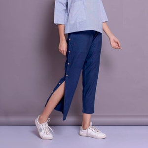 May include: A woman is wearing a blue and white striped top with a pocket and blue pants with button detailing. She is wearing white sneakers.