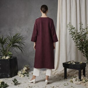 May include: A burgundy-colored linen dress with a long, flowing silhouette and three-quarter sleeves. The dress features a simple, round neckline and side slits. The model is wearing white pants and shoes.