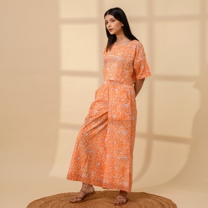 May include: An orange two-piece outfit with a floral pattern. The set includes a short-sleeved top and wide-leg pants. The outfit is made of a lightweight fabric and is suitable for warm weather. The model is wearing brown sandals.