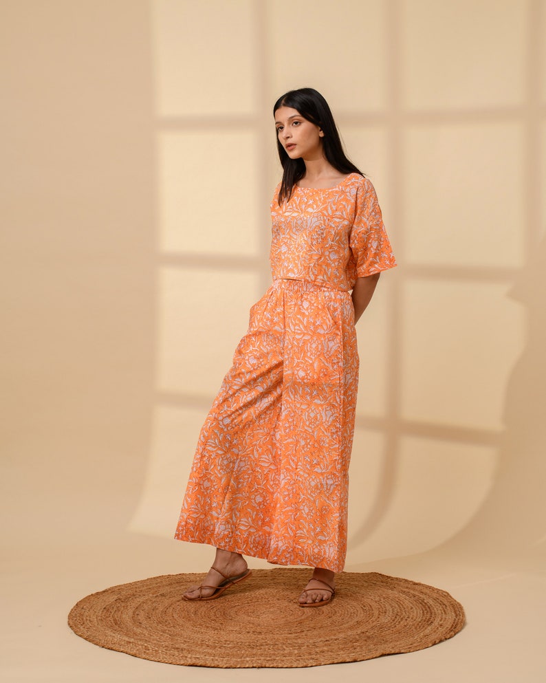 May include: An orange two-piece outfit with a floral pattern. The set includes a short-sleeved top and wide-leg pants. The outfit is made of a lightweight fabric and is suitable for warm weather. The model is wearing brown sandals.