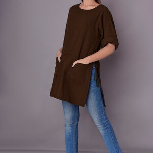 May include: A brown, long-sleeved tunic with side slits and a pocket on the front. The tunic is worn with blue jeans and white sandals.