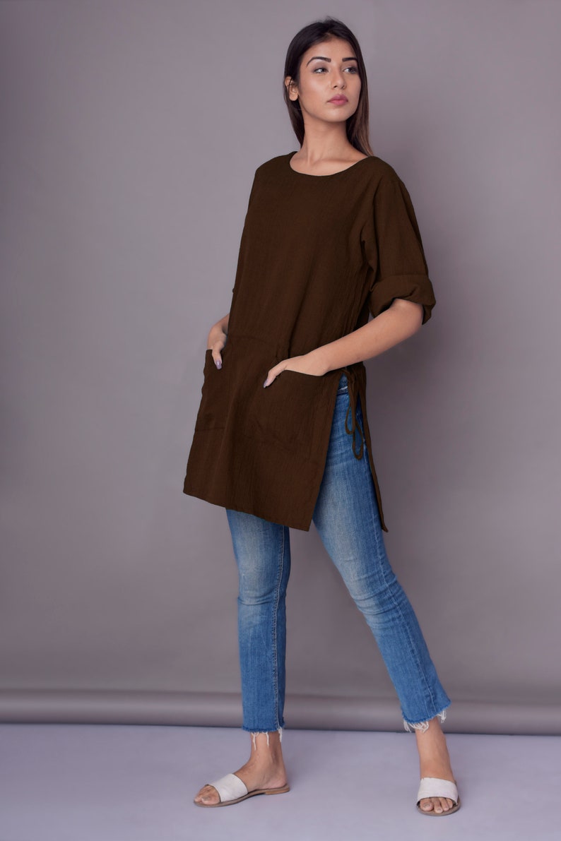May include: A brown, long-sleeved tunic with side slits and a pocket on the front. The tunic is worn with blue jeans and white sandals.
