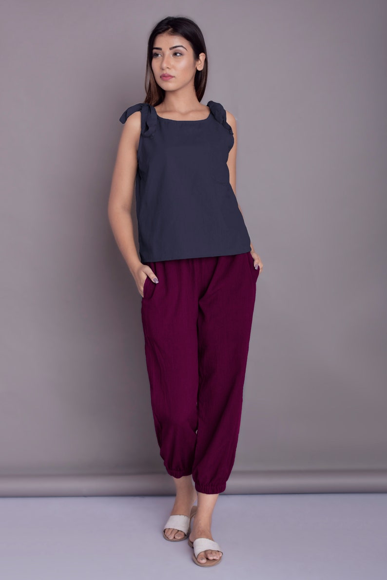 May include: A woman is wearing a dark blue sleeveless top with ruffled straps and maroon trousers. She is standing on a light grey background.
