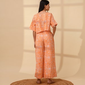 May include: An orange two-piece outfit with a floral pattern. The set includes a short-sleeved crop top and wide-leg pants. The outfit is made of a lightweight fabric and is suitable for warm weather. The model is standing on a round, woven rug.