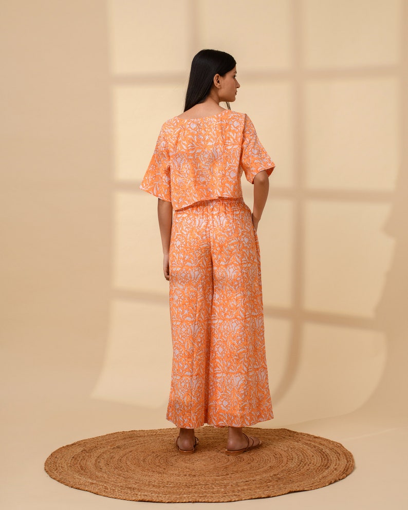 May include: An orange two-piece outfit with a floral pattern. The set includes a short-sleeved crop top and wide-leg pants. The outfit is made of a lightweight fabric and is suitable for warm weather. The model is standing on a round, woven rug.
