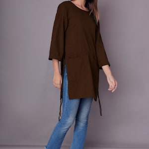May include: A brown long-sleeved tunic with side slits and tie closures. The tunic is worn with blue jeans and white sandals.