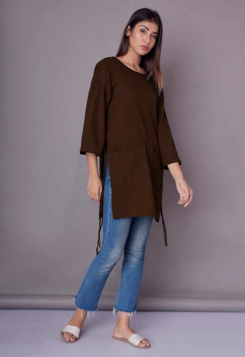 May include: A brown long-sleeved tunic with side slits and tie closures. The tunic is worn with blue jeans and white sandals.