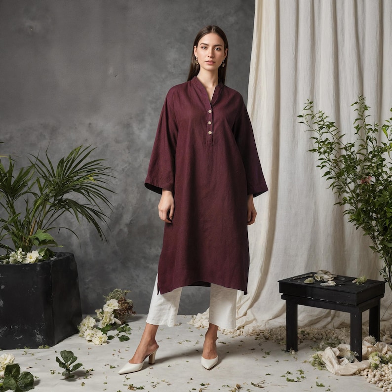 May include: A burgundy tunic dress with a mandarin collar and three visible buttons. The dress has long sleeves and is paired with white cropped pants and white heels. The background features plants and a small black table.