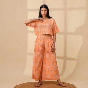 May include: An orange two-piece outfit featuring a crop top and wide-leg pants. The set has a white floral pattern. The model is wearing brown sandals and standing on a round, brown rug. The outfit is made of a lightweight fabric.