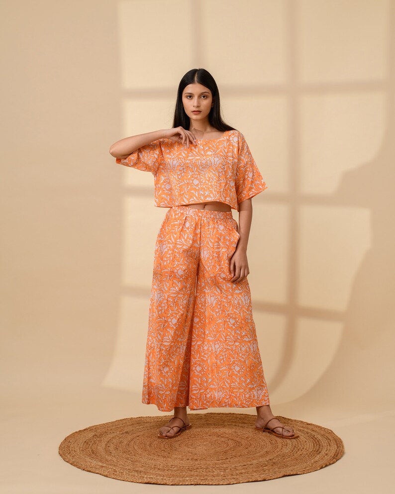 May include: An orange two-piece outfit featuring a crop top and wide-leg pants. The set has a white floral pattern. The model is wearing brown sandals and standing on a round, brown rug. The outfit is made of a lightweight fabric.