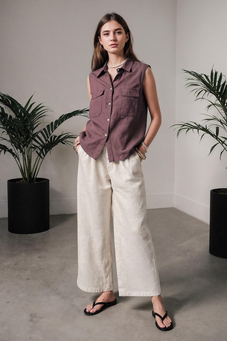 May include: A sleeveless, button-down shirt in a muted purple hue, paired with wide-leg, cream-colored pants. The outfit is completed with black flip-flops. The model is wearing a pearl necklace and bracelets. The setting includes potted plants.