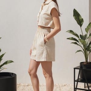 May include: A beige linen two-piece set, consisting of a cropped button-down shirt and high-waisted shorts. The shorts have pockets. The model is wearing orange sandals. The outfit is suitable for warm weather.