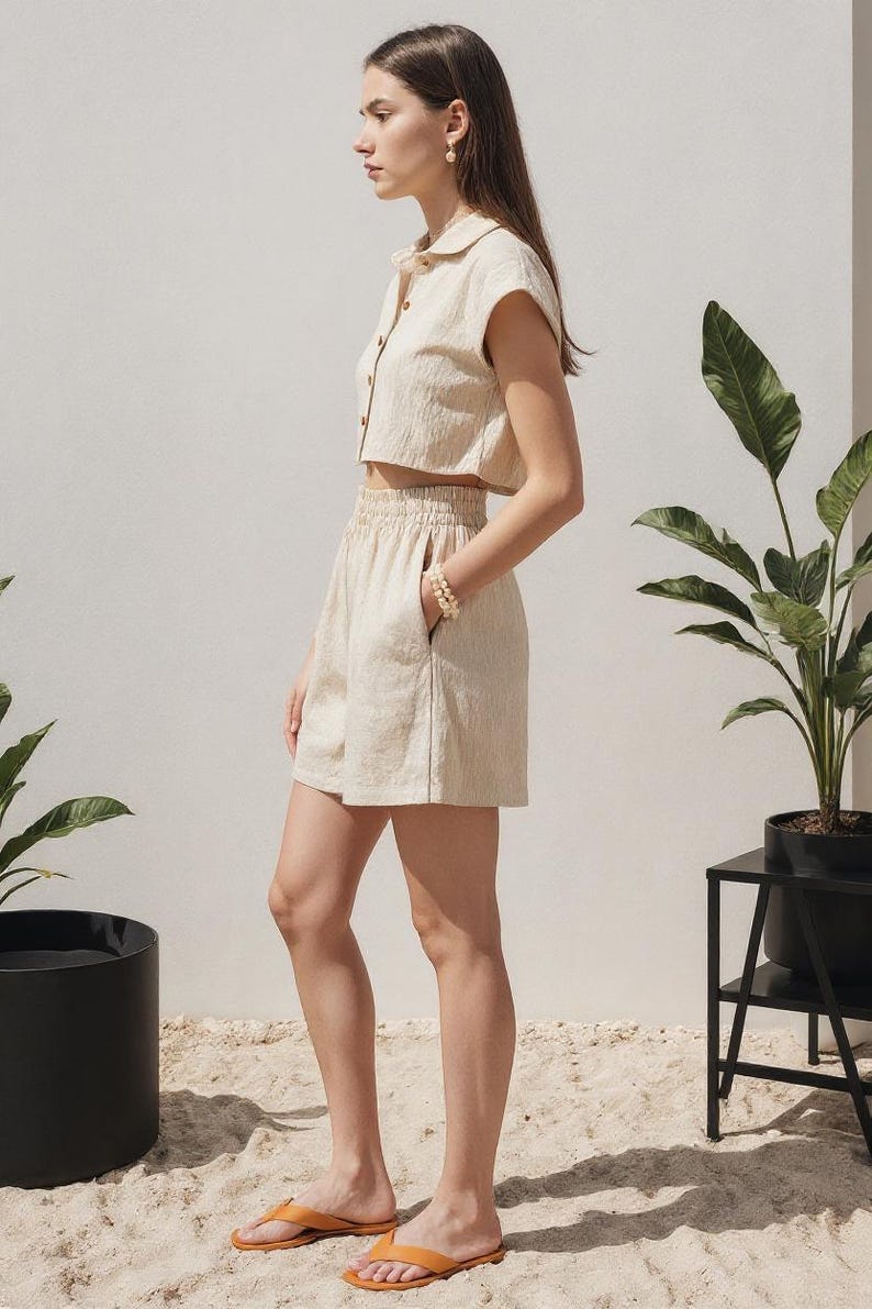 May include: A beige linen two-piece set, consisting of a cropped button-down shirt and high-waisted shorts. The shorts have pockets. The model is wearing orange sandals. The outfit is suitable for warm weather.