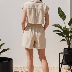May include: A beige linen shorts set, featuring a cropped collared shirt and high-waisted shorts. The outfit is made of a lightweight fabric. The model is wearing orange sandals. The background includes plants and a white wall.