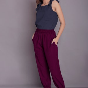 May include: A woman is wearing a dark blue sleeveless top with a tie detail at the shoulders and burgundy trousers with an elastic waistband. She is standing on a light grey surface.