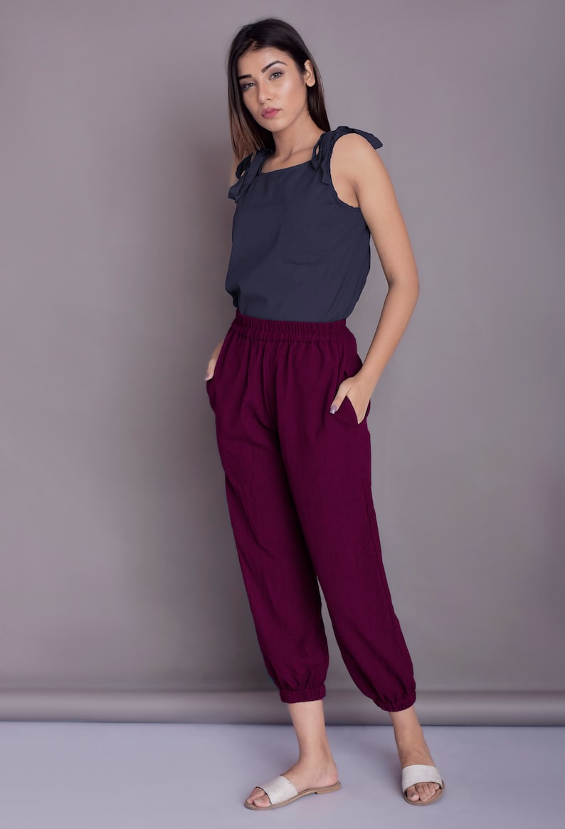 May include: A woman is wearing a dark blue sleeveless top with a tie detail at the shoulders and burgundy trousers with an elastic waistband. She is standing on a light grey surface.